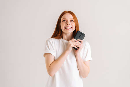 Studio portrait of happy dreaming young woman with wide smile reading good online message using mobile phone on white isolated background. Cheerful lady holding smartphone in hands and looking up.の写真素材