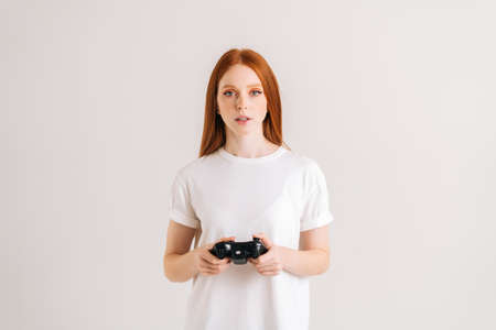 Studio portrait of attractive young woman playing video game with controller looking at camera on white isolated background. Happy lady using joystick on console enjoying online game.の写真素材