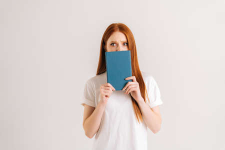 Studio portrait of scared young student woman hides face behind notebook looking at camera, standing on white isolated background. Astonished redhead lady posing with educational materials.の写真素材