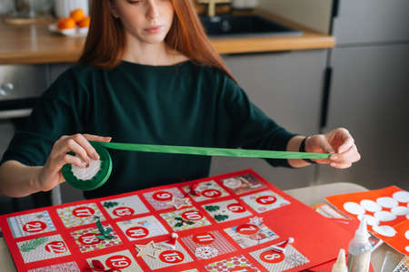 Cropped shot of focused redhead young woman using holiday ribbon to frame board with advent calendar envelopes at home. Concept of preparing for xmas, new year and winter holidays, festive mood.の写真素材