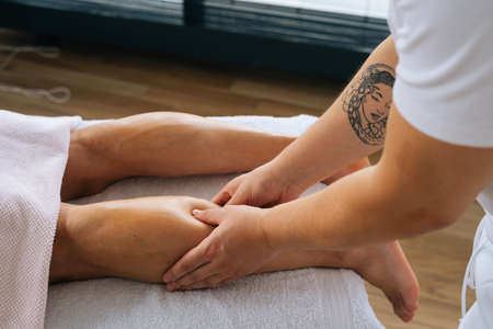 Close-up side view hands of unrecognizable male masseur with strong tattooed hands massaging lower leg of muscular athlete man after physical sports workout, lying on stomach at massage table.の写真素材