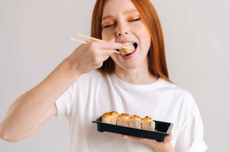 Close-up face of happy young woman with closed eyes eating delicious sushi rolls with chopsticks standing on white isolated background in studio. Pretty Caucasian redhead female chewing Asian food.の写真素材