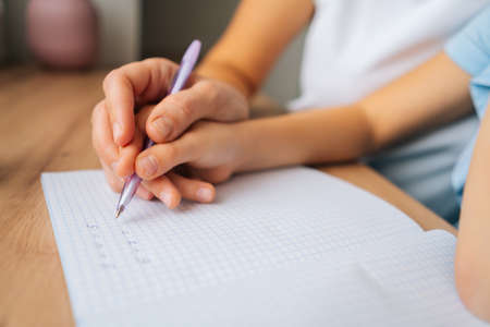 Close-up cropped shot of primary daughter doing homework writing in copybook with pen with young mother sitting at home table. Side view of female tutor teaching elementary girl helping with lesson.の写真素材