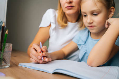 Close-up cropped shot of primary daughter doing homework with young mother sitting at home table by...