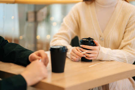 Close-up of woman and man sitting in cafe holding warm cups of coffee on table having fun conversation. Young couple in love enjoying time spending with each other sitting at table in cozy coffee shopの写真素材