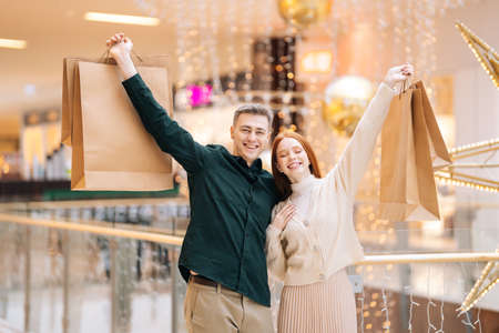 Portrait of happy beautiful young couple in love holding shopping bags standing in mall and looking at camera. Joyful handsome man and attractive woman purchasing together at store center.の写真素材