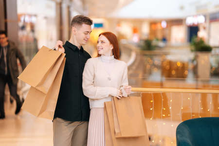 Portrait of smiling pretty young couple holding shopping paper bags with purchases and looking at...