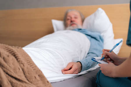 Close-up of unrecognizable female doctor writing down diagnosis and symptoms in medical history on clipboard, talking to sick senior male patient lying on bed in hospital, selective focus.の写真素材