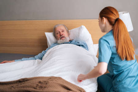 Unrecognizable young female nurse helping senior adult male patient to lying on bed and covering him blanket at home. Friendly woman taking care of mature old patient and talking to himの写真素材