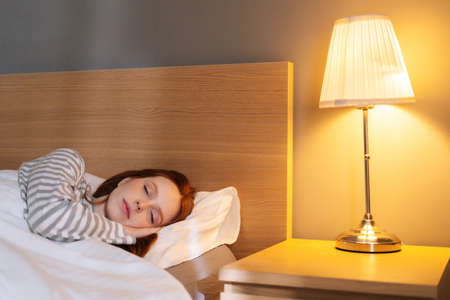 Close-up face of tired young woman sleeping well in bed hugging soft white pillow at home, bedside lamp lighting with warm yellow light. Cute lady resting enjoying fresh soft bedding linen in bedroomの写真素材