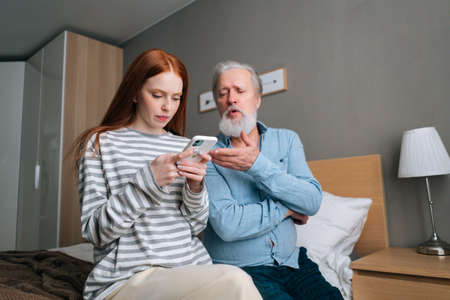 Low-angle view of pretty granddaughter showing photo on mobile photo to gray-haired grandfather sitting on bed at home. Attractive young woman teaching mature adult male how using smartphone.の写真素材
