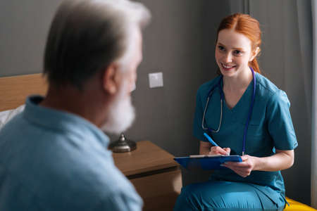Close-up view from back of senior male patient talking with smiling female doctor writing down diagnosis and symptoms in medical history on clipboard at nursing home, selective focus.の写真素材