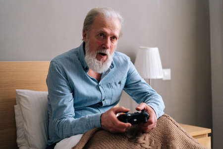Front view of focused gray-haired senior male playing online video game using gamepad at home. Happy bearded mature elderly man in pajama playing computer game having fun with joysticks.の写真素材