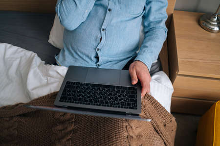 Close-up top view of unrecognizable mature adult male using typing on laptop computer sitting on bed with blanket at home in morning. Senior older business man chatting online with colleagues.の写真素材