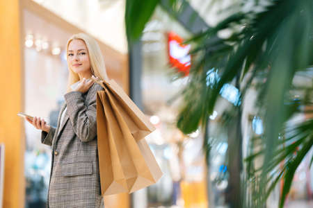 Low-angle view of elegance attractive blonde young woman in stylish coat looking at camera holding mobile phone and shopping paper bags with purchases in hall of mall centre, blurred background.の写真素材