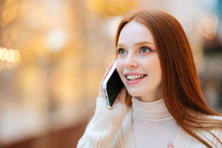 Close-up face of happy attractive redhead young woman talking on mobile phone standing in shopping mall with bright interior during holiday sales, looking away, blurred background, bokeh lights.の写真素材