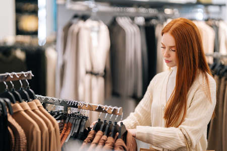 Side view of serious pretty female shopaholic choosing clothes from rack in clothing store, blurred background. Cute young woman shopper select and buying clothes in fashion boutique during sale.の写真素材