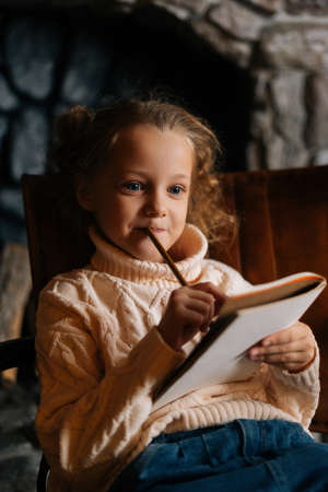 Close-up vertical shot of dreaming little blonde curly child girl writing letter to Santa Claus sitting on chair at home by fireplace. Pretty Caucasian kid writes letter with wishes for gift.の写真素材
