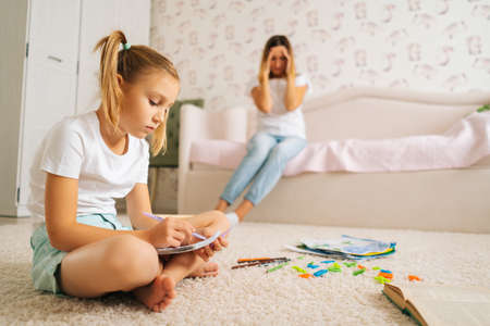 Side view of primary girl child writing homework in copybook sitting on floor with laptop for distance education online at home, on blurred background of stressed young mother sitting on sofa.の写真素材
