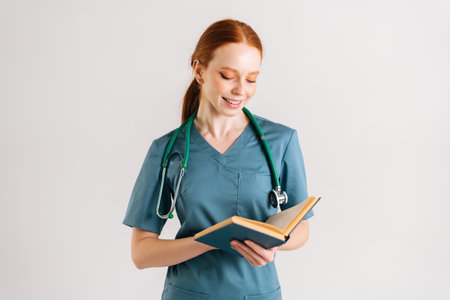 Portrait of positive young woman physician in green uniform with stethoscope reading medical book standing on white isolated background in studio. Female doctor reading paper treatise.の写真素材