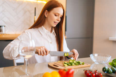 Close-up side view of attractive young redhead woman cutting fresh cucumber cooking food salad...
