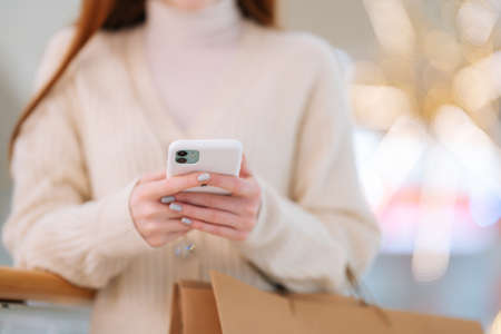 Close-up cropped shot of unrecognizable female shopaholic using typing smartphone holding shopping...