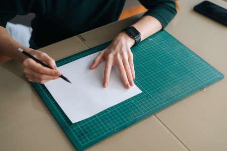 Close-up top view of unrecognizable female designer writing making mark with pencil on white blank paper lying on green rubber cutting mat at desk, selective focus. Concept of creative work and hobbyの写真素材