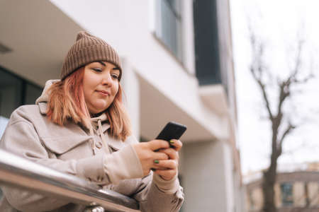 Close-up low-angle view of overweight woman in autumn hat typing online message, browsing social medial using mobile phone standing in city street on background of office building in cloudy dayの写真素材