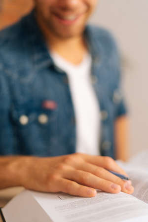 Vertical selective focus shot of happy young man reading paper book holding pen in hands sitting at desk in home office. Handsome student male studying alone with textbook, blurred background.の写真素材