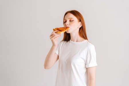 Studio portrait of happy attractive young woman with closed eyes appetite eating delicious pizza standing on white isolated background. Pretty redhead female eating tasty meal.の写真素材