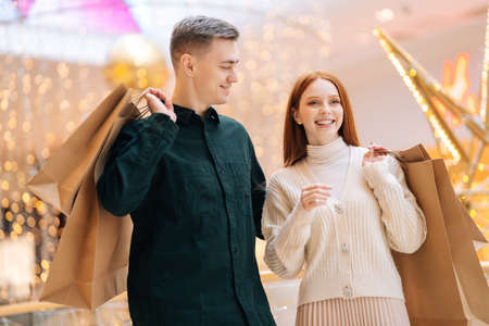 Portrait of cheerful beautiful young couple holding shopping bags in shoulders walking in mall,...