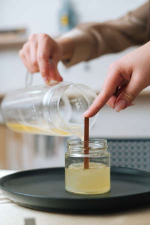 Close-up vertical shot of unrecognizable female artisan pouring melted wax into tranparent glass jar...