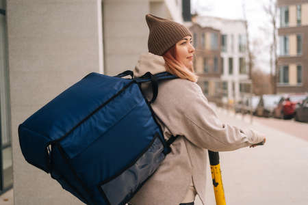 Back view of female courier food delivery with big thermal backpack walking with electric scooter in city street. Adult delivery woman in warm hat and jacket delivering order to client.の写真素材
