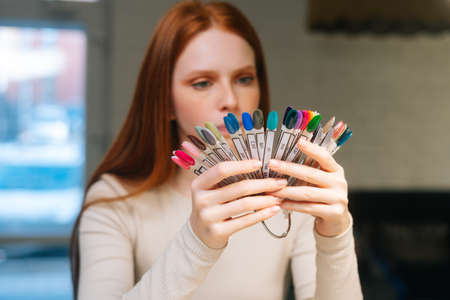 Close-up face of thoughtful pretty young woman client choosing colour from colourful nails tips in...