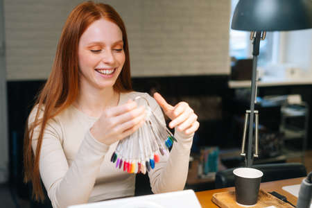 Portrait of cheerful pretty young woman client choosing colour from colourful nails tips in manicure...