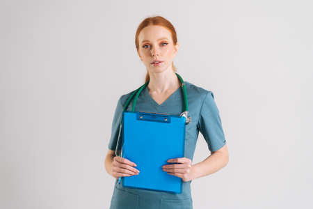 Portrait of serious young female physician in medical uniform holding clipboard and looking at camera, standing on white isolated background in studio. Front view of confident nurse with stethoscope.の写真素材