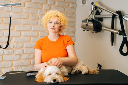Portrait of female groomer stroking curly Labradoodle dog before brushing and shearing, preparation to bathing at grooming salon, looking at camera. Woman pet hairdresser doing professional care.の写真素材