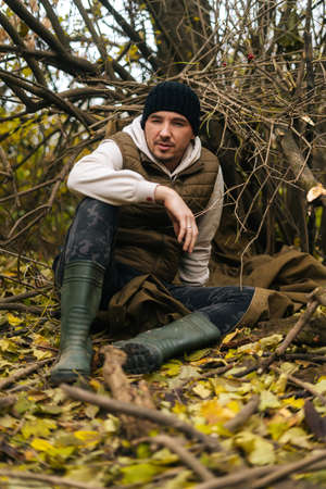 Vertical shot of tired tourist man sitting on spread out raincoat in thicket under tree in woods on...