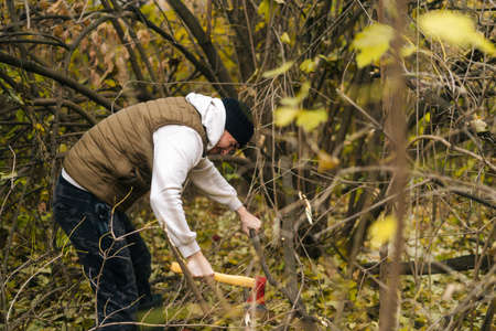 Strong tourist male wearing warm clothes chopping firewood with axe in forest on overcast cold day. Concept of bushcraft, camping and survival in nature.の写真素材