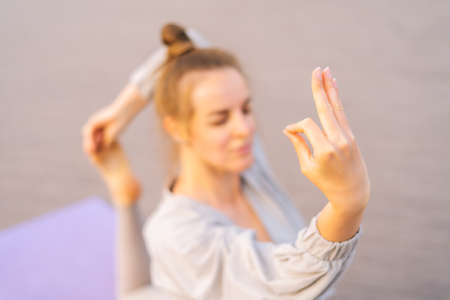 Blurred shot of flexible Caucasian young yogini woman in sportswear performing yoga Pigeon pose outdoors sitting on yoga mat. Sporty calm female stretching in city park at cloudy summer day outside.の写真素材
