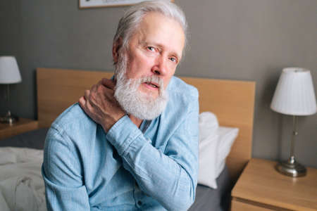 Close-up of senior adult man warming up and massaging shoulder sitting on bed in morning after waking up at home, looking at camera. Bearded mature man suffering from muscle tension after waking up.の写真素材