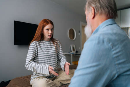 Pretty young woman and mature male having fun, enjoying leisure time, sharing news at home. Back view of unrecognizable bearded grandfather talking with pretty young granddaughter sitting in bed.の写真素材