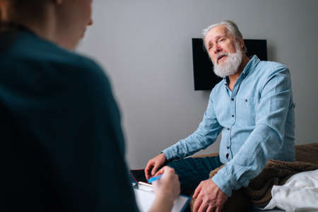 Close-up view from back of female doctor talking to illness senior male patient sitting on bed at nursing home, writing down diagnosis and symptoms in medical history on clipboard, selective focus.の写真素材