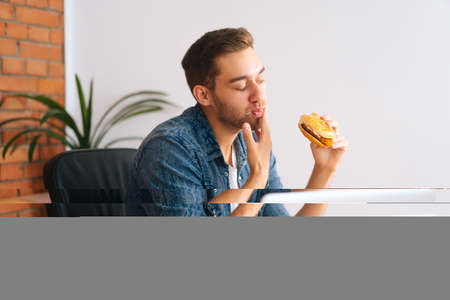 Hungry freelance programmer male eating hamburger watching online video on laptop computer during...