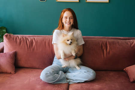 Portrait of happy smiling young woman sitting on comfortable sofa cuddling pretty small white spitz...