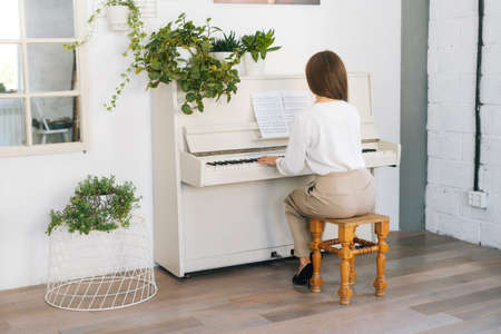 Back view of unrecognizable young woman pianist playing on white classical piano in light classroom....
