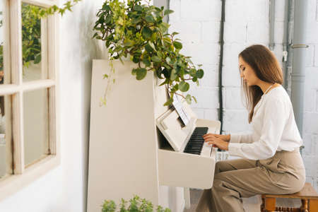 Side view of focused young female musician playing on white classical piano energetic music at home studio during lesson. Lady practicing piano lesson looking on musical notes in classroom.の写真素材