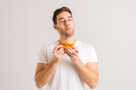 Portrait of handome young man with enjoying eating delicious slice of pizza, with closed eyes from pleasure on white isolated background. Studio shot of hungry male student eating tasty food.の写真素材