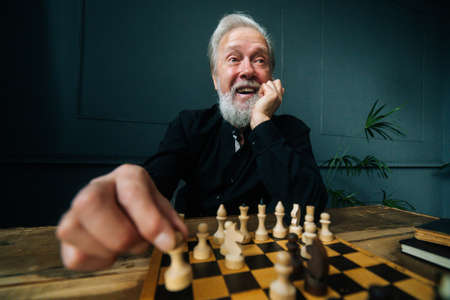Close-up front view of cheerful bearded mature male performing move with pawn piece on wooden...
