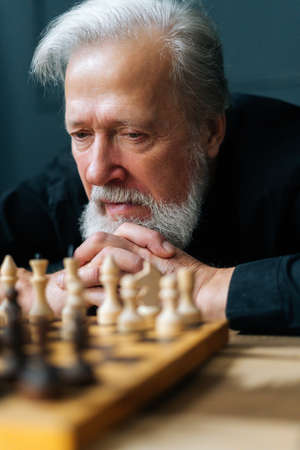 Vertical close-up shot of thoughtful gray-haired senior male thinking game strategy sitting on...